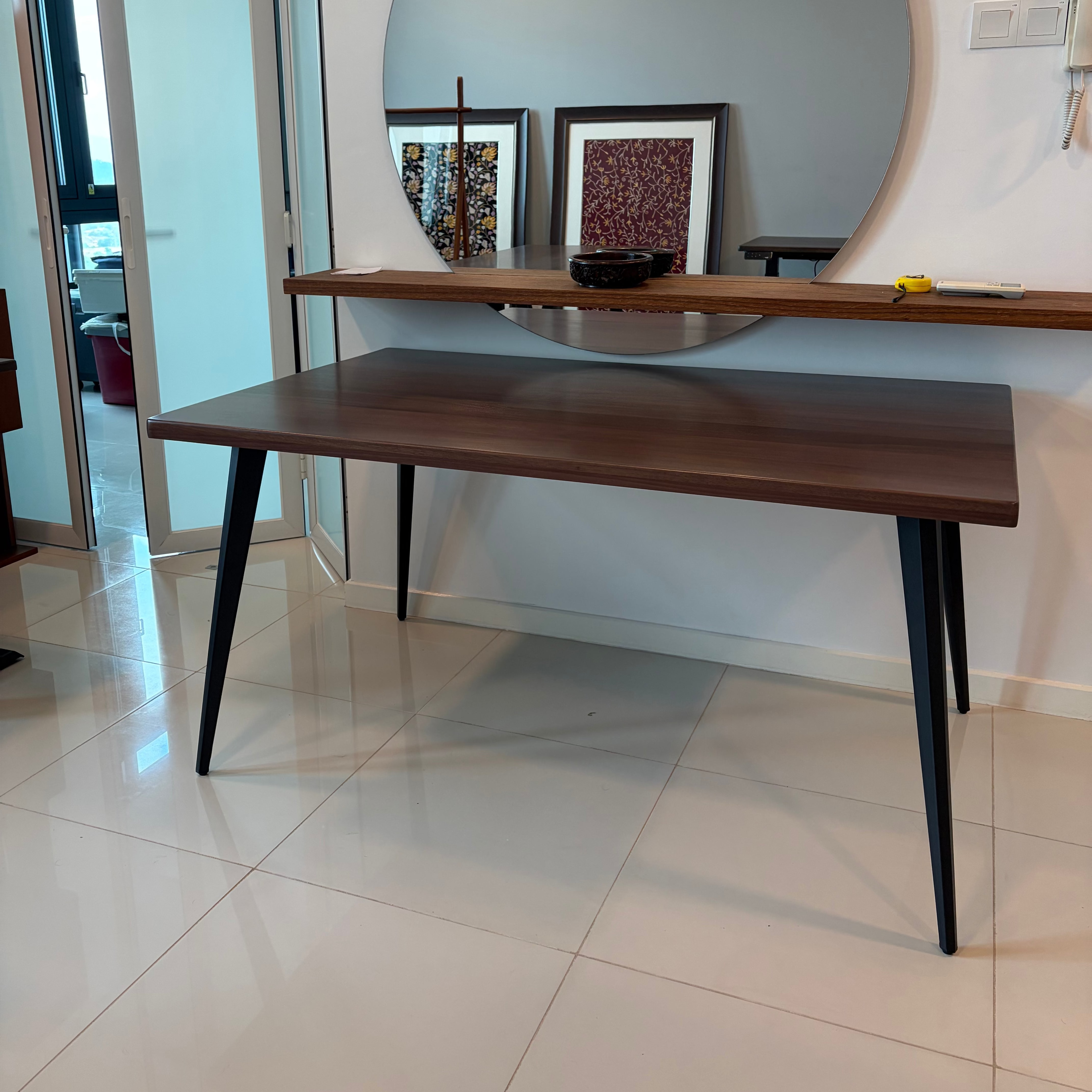 Wooden dining table with black legs in a room with a large round mirror on the wall.
