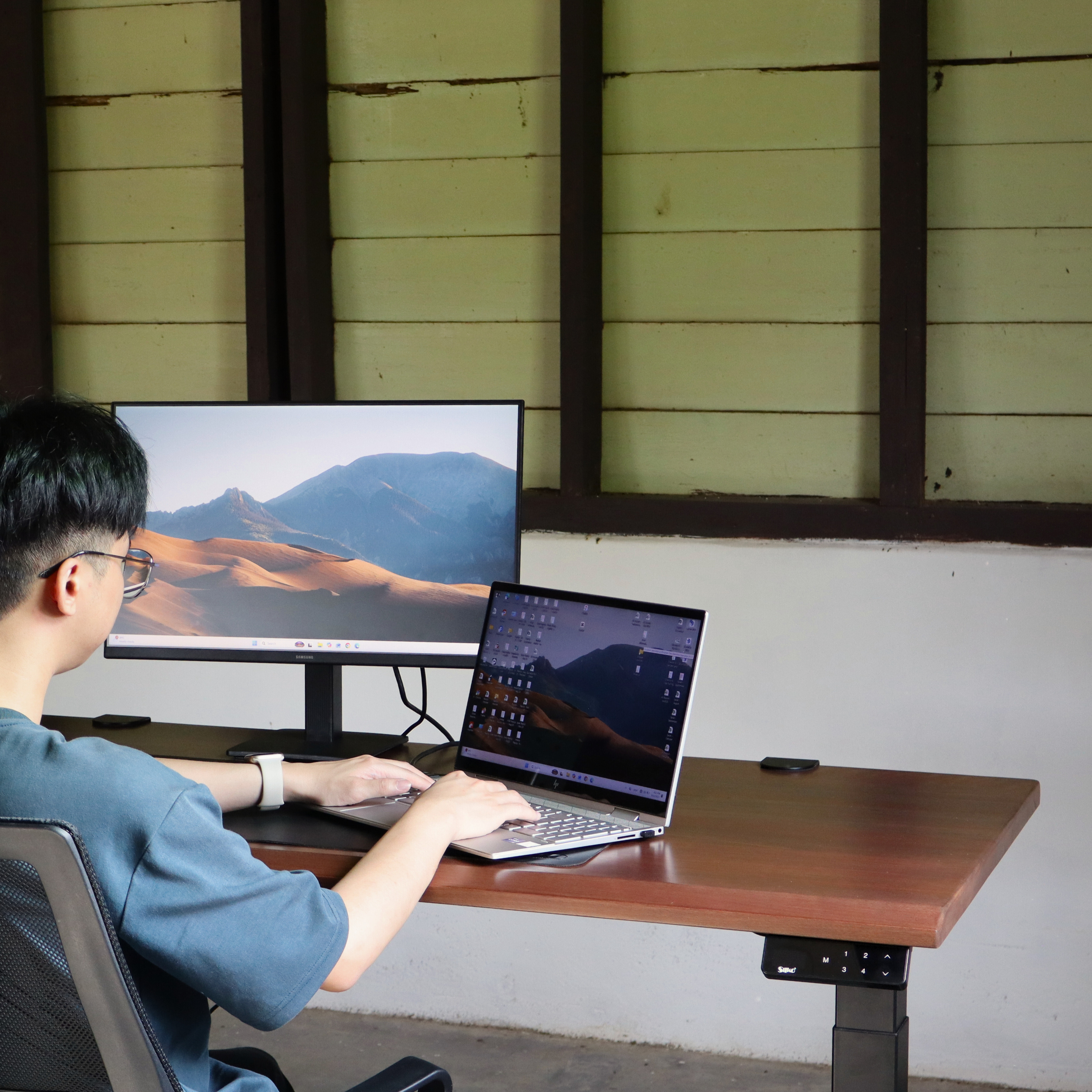 Person using a laptop and monitor at a wooden standing desk with a mountainous landscape on screens.