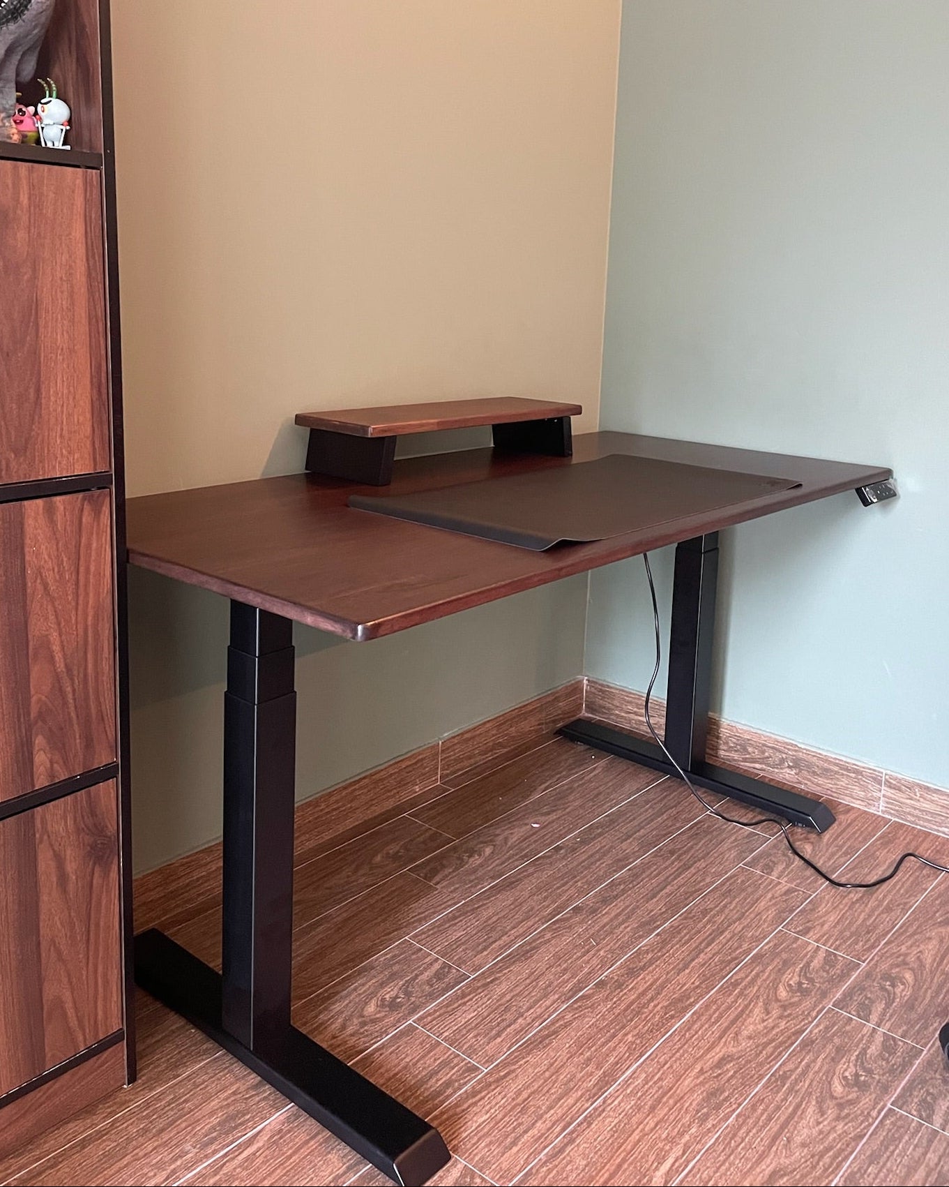 Wooden desk with adjustable height in a room with a bookshelf and plain walls.