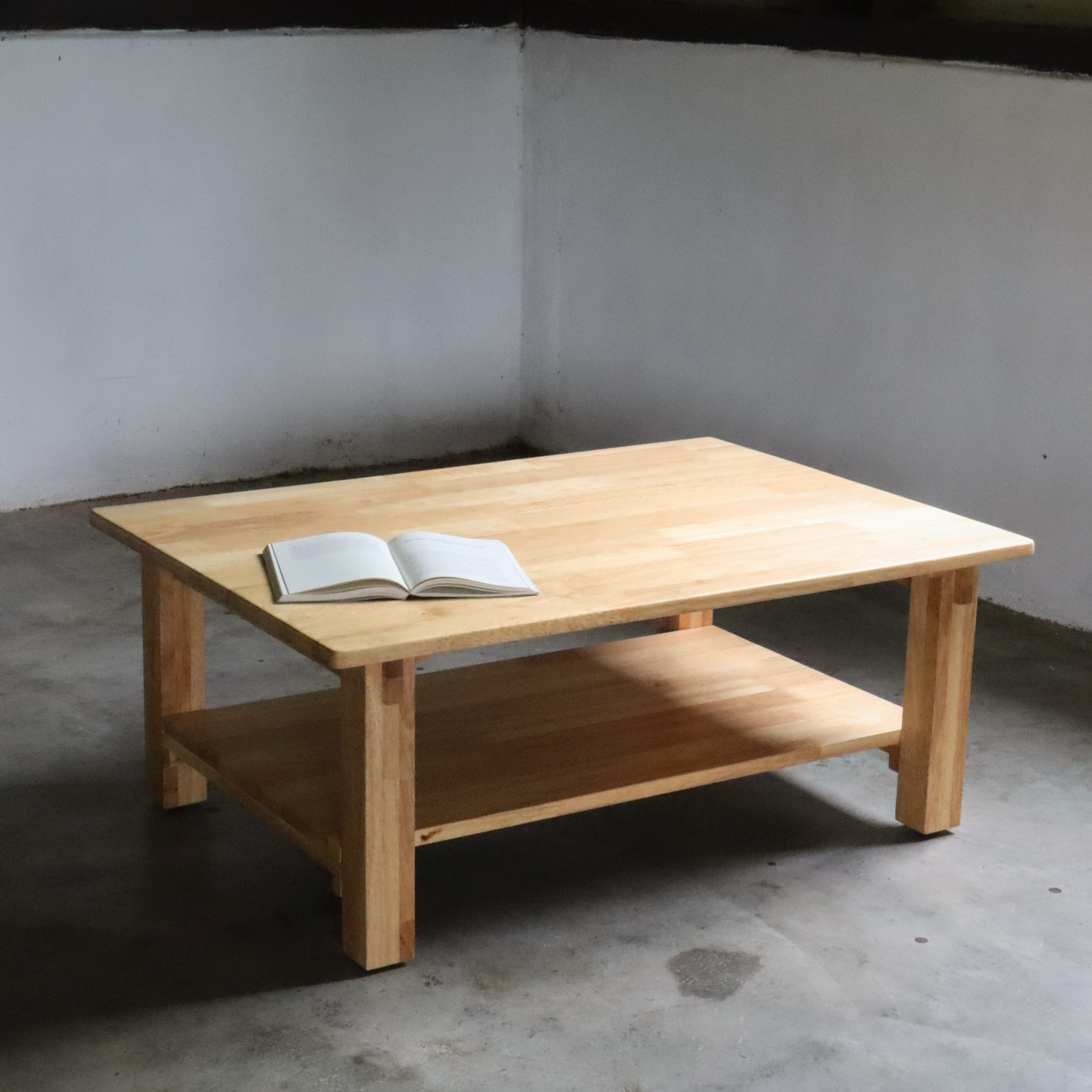 Wooden coffee table with an open book on a concrete floor against a plain wall.