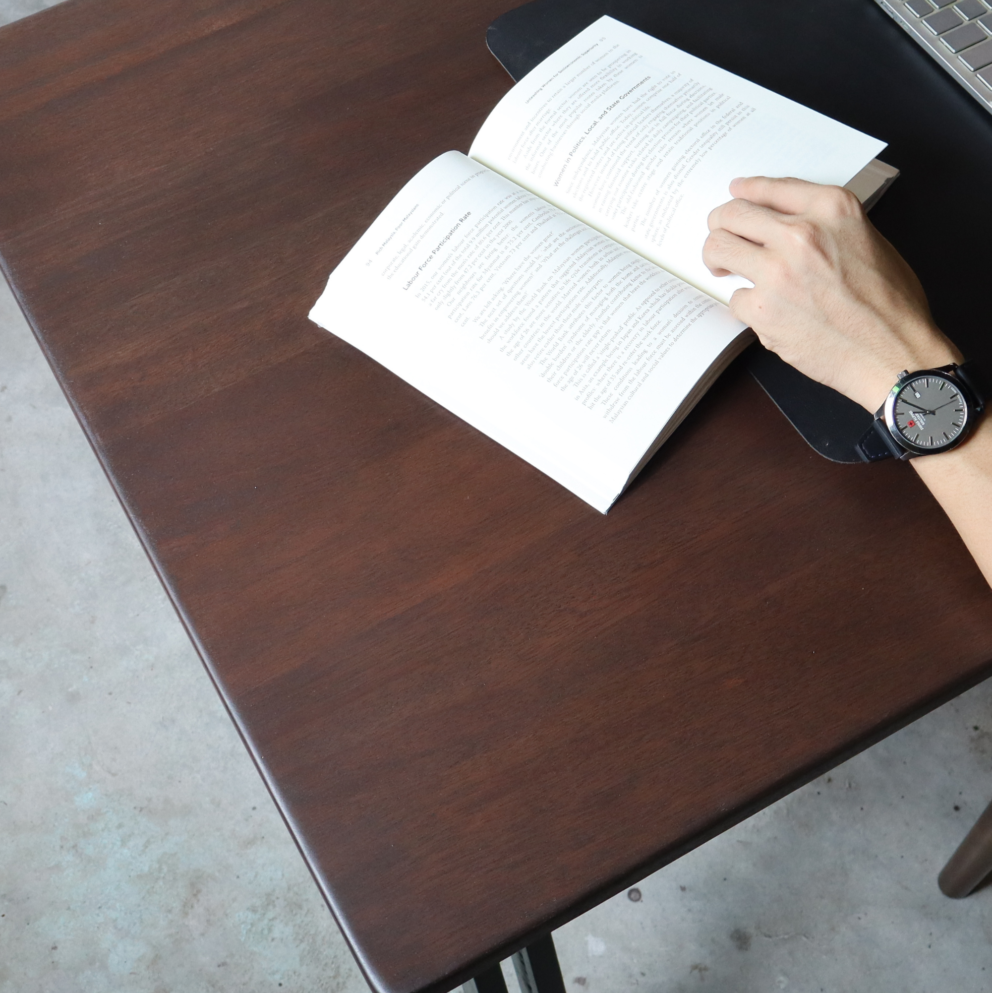 Person's hand holding a pen over an open book on a wooden desk with a laptop in the background.