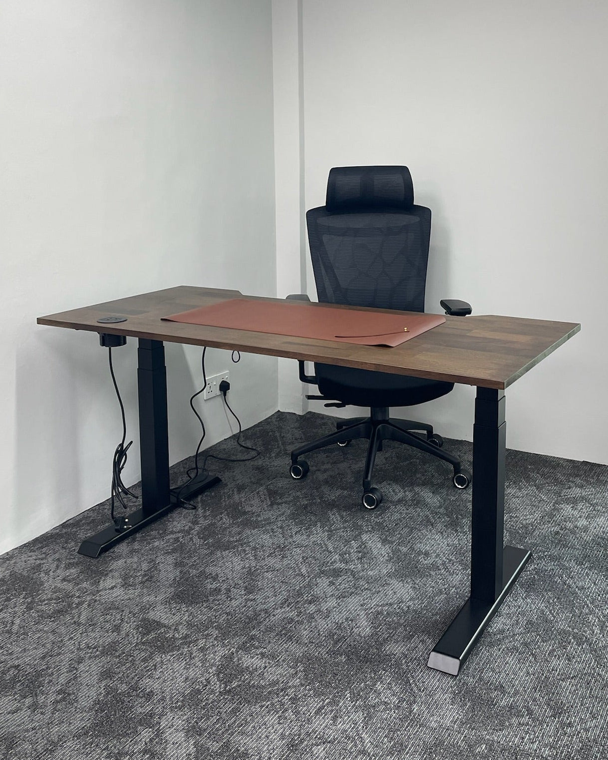 Wooden standing desk with black chair in a room with gray carpet and white walls.