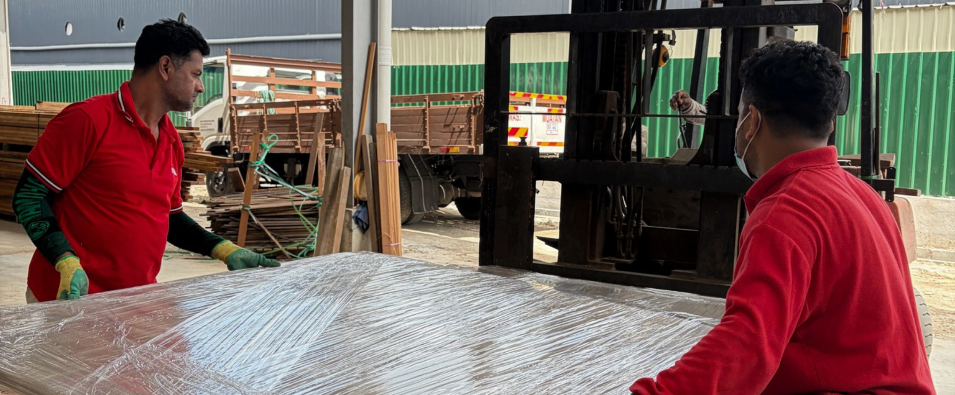 Two workers in red shirts handling a large sheet of material with a forklift in the background.