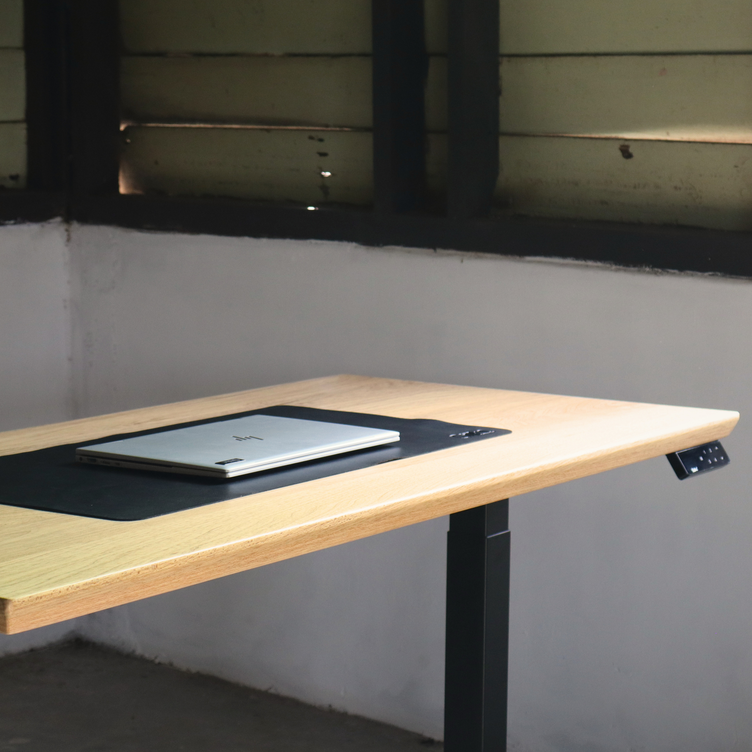 Wooden standing desk with a laptop on a black mouse pad against a plain wall.